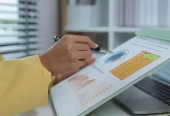Imagem de uma pessoa analisando gráficos e dados em um caderno enquanto utiliza um laptop ao fundo, representando a importância da análise de dados para tomada de decisões