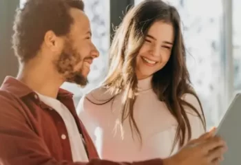 Um casal sorrindo enquanto observa um tablet em um ambiente iluminado. Eles parecem felizes e engajados, sugerindo interações de customer care.