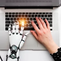Imagem de uma mão humana e uma mão robótica digitando em um laptop, representando a integração entre humanos e inteligência artificial no uso de tecnologia.