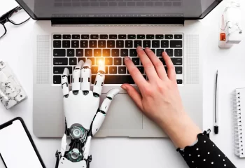 Imagem de uma mão humana e uma mão robótica digitando em um laptop, representando a integração entre humanos e inteligência artificial no uso de tecnologia.