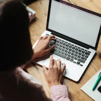 Mulher com camisa rosa digitando em laptop prateado sobre mesa de madeira com caderno, caneta e celular ao lado, ilustrando conteúdo da Orgânica sobre conteúdo orgânico.