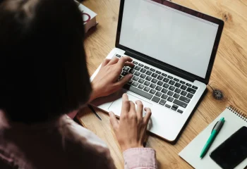 Mulher com camisa rosa digitando em laptop prateado sobre mesa de madeira com caderno, caneta e celular ao lado, ilustrando conte&uacute;do da Org&acirc;nica sobre conte&uacute;do org&acirc;nico.