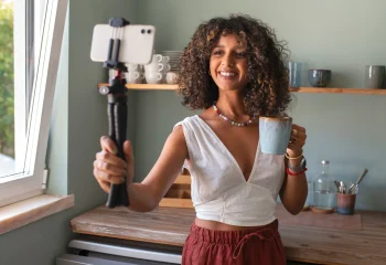 Mulher segurando uma caneca com uma das m&atilde;os, enquanto segura um estabilizador com um celular na outra, gravando um conte&uacute;do UGC, ilustrando um artigo de mesmo tema para o blog da Org&acirc;nica.