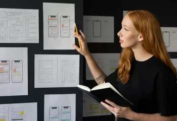 Designers analisando protótipos de aplicativos móveis para desenvolvimento de produto em uma parede com sketches e wireframes.