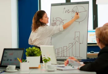 Mulher apresentando gr&aacute;ficos de crescimento e diagramas em quadro branco durante reuni&atilde;o, ilustrando um conte&uacute;do da Org&acirc;nica sobre estrat&eacute;gia de conte&uacute;do.