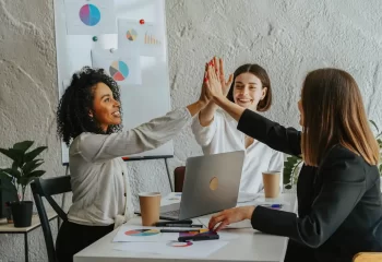 Três mulheres de escritório comemorando uma conquista, símbolo de sucesso em estratégias de go-to-market, em ambiente de trabalho colaborativo com gráficos.