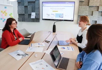Reuni&atilde;o de equipe em escrit&oacute;rio moderno com tr&ecirc;s mulheres discutindo estrat&eacute;gias de marketing digital, ilustrando um conte&uacute;do da Org&acirc;nica sobre ideias de conte&uacute;do.