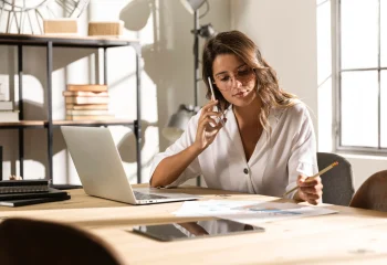 Mulher trabalhando em escrit&oacute;rio moderno com notebook, falando ao telefone enquanto faz anota&ccedil;&otilde;es em um papel, ilustrando um conte&uacute;do da Org&acirc;nica sobre lead frio, morno e quente.