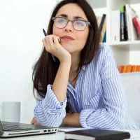 Mulher jovem pensativa segurando caneta perto do rosto em ambiente de escritório com laptop e estante de livros ao fundo, ilustrando um artigo no blog da Orgânica sobre lead magnet.