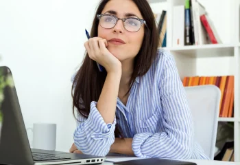 Mulher jovem pensativa segurando caneta perto do rosto em ambiente de escrit&oacute;rio com laptop e estante de livros ao fundo, ilustrando um artigo no blog da Org&acirc;nica sobre lead magnet.