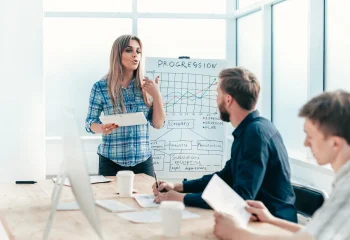 Mulher apresentando gr&aacute;fico de progresso em reuni&atilde;o de neg&oacute;cios com colegas, ilustrando um conte&uacute;do da Org&acirc;nica sobre lista de leads.