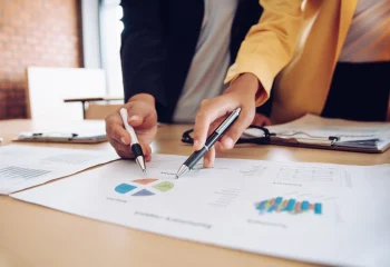 Duas pessoas analisando gr&aacute;ficos e relat&oacute;rios em uma mesa de escrit&oacute;rio, ilustrando um conte&uacute;do da Org&acirc;nica sobre marketing estrat&eacute;gico.