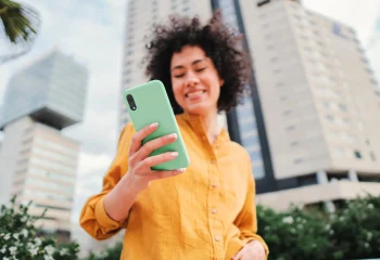 Mulher sorrindo usando camisa amarela segurando smartphone com capa verde em área urbana com prédios ao fundo