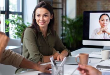 Mulher sorrindo em uma reunião no escritório com colegas, enquanto uma videoconferência ocorre em um monitor ao fundo.