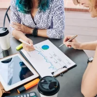 Duas mulheres analisando gráficos financeiros e dados em papel com tablet, smartphone e café em mesa redonda